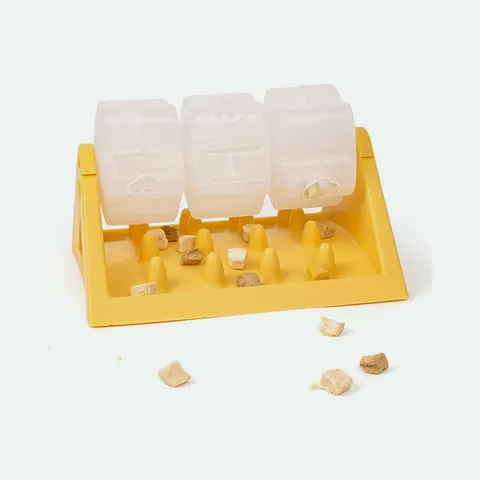 Yellow rotating treat feeder with three clear drums on a triangular stand, kibble scattered on the ridged base